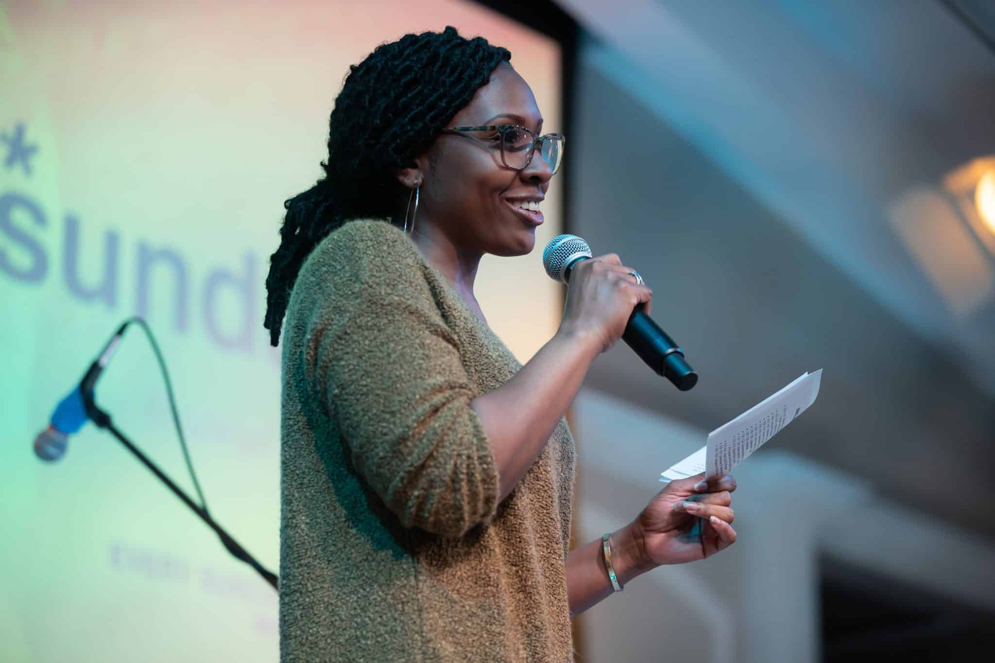 A woman wearing eyeglass and smile talking o the microphone